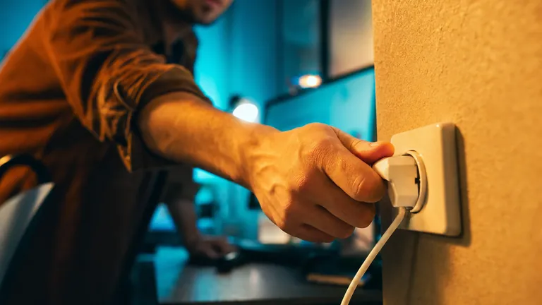 A hand pulls a power plug out of a socket in front of a computer workstation