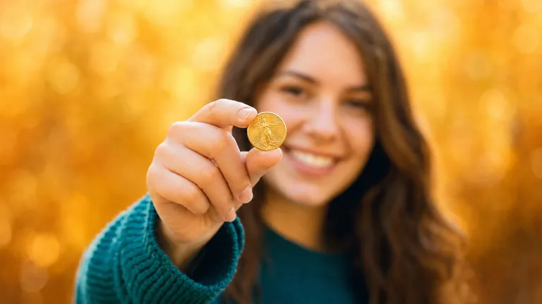 A smiling young woman in a teal sweater holds a small coin out toward the camera